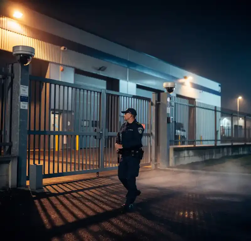 Beveiliger in uniform patrouilleert 's nachts bij een industrieel pand met stalen poort, CCTV-camera's en mistsfeer.